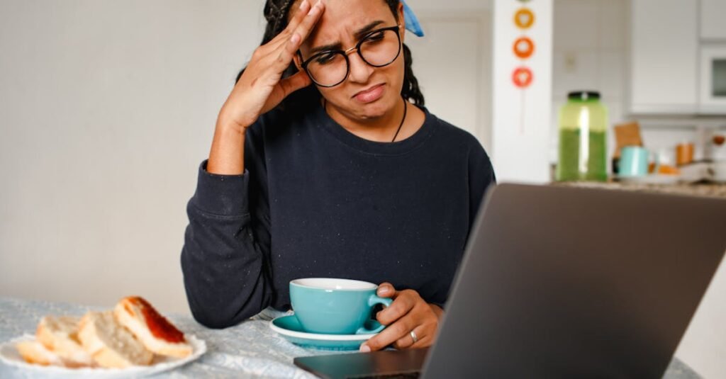 A woman holding her head in frustration while working on a laptop at home, drinking coffee.
