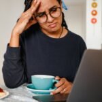 Dificuldade de concentração: 7 causas físicas comuns 7 A woman holding her head in frustration while working on a laptop at home, drinking coffee.