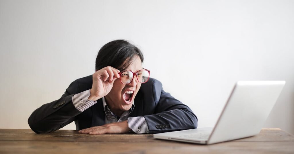 Cansaço constante: 9 causas que deve conhecer 11 Modern Asian man in jacket and glasses looking at laptop and screaming with mouth wide opened on white background