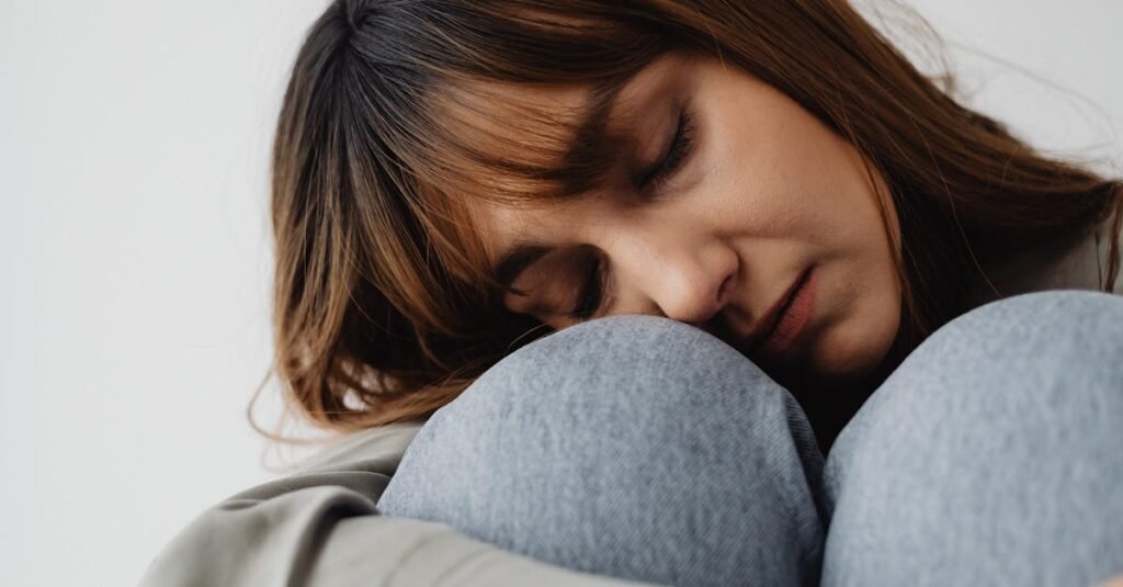 Mulher com expressão de exaustão, apoiada nos joelhos e de olhos fechados, ilustrando cansaço constante e necessidade de descanso.