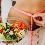 9 mitos sobre nutrição em que você ainda acredita 4 Mulher a medir a cintura com fita métrica enquanto segura prato com salada, ilustrando mitos sobre nutrição e controlo alimentar