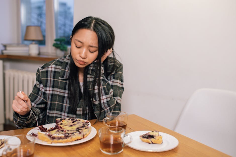 5 sinais de fome emocional que confundem o corpo Mulher pensativa a comer bolo à mesa ilustrando sinais de fome emocional que confundem o corpo e relação entre emoções e alimentação Vida em Equilíbrio