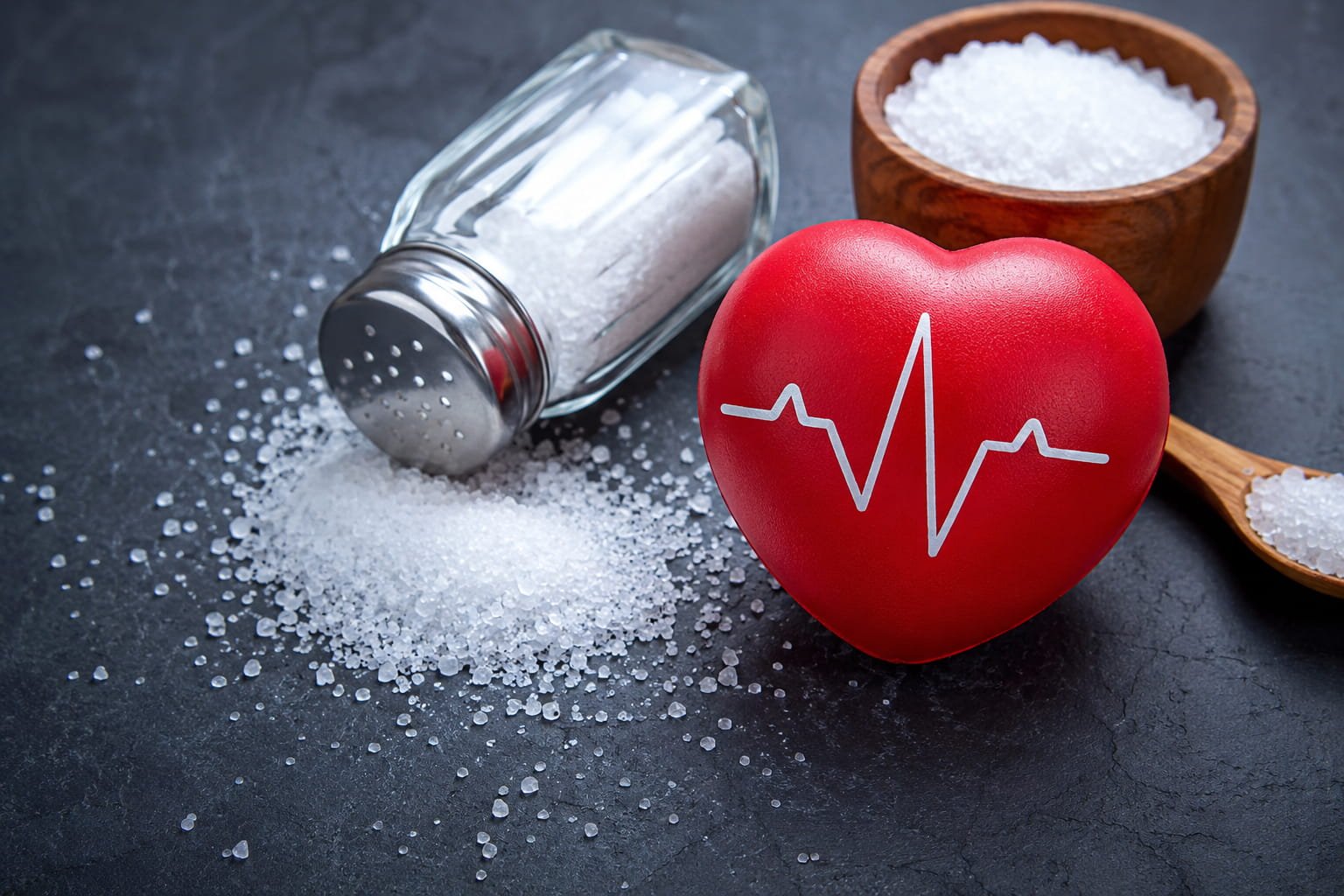 Coração vermelho ao lado de sal derramado e saleiro, simbolizando o impacto do excesso de sal na saúde cardiovascular